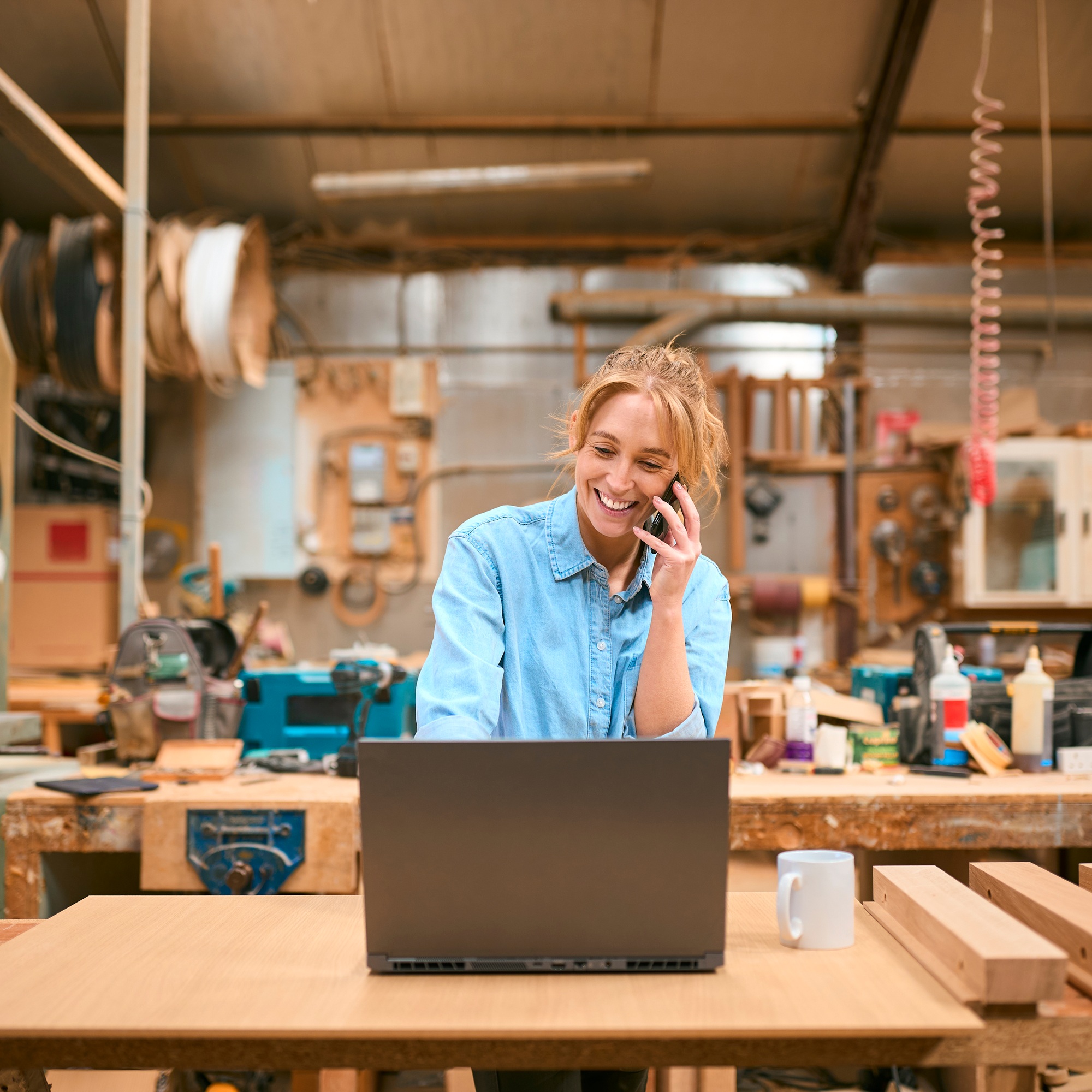 Female Carpenter Working In Woodwork Workshop Talking On Mobile Phone And Using Laptop